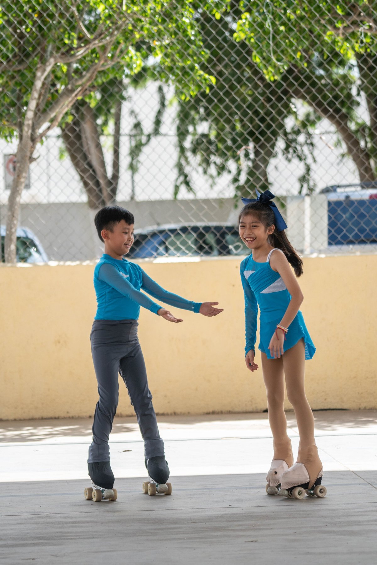 Dos niños con camisetas azules patinando juntos en un patio exterior soleado con árboles y una reja de alambre de fondo