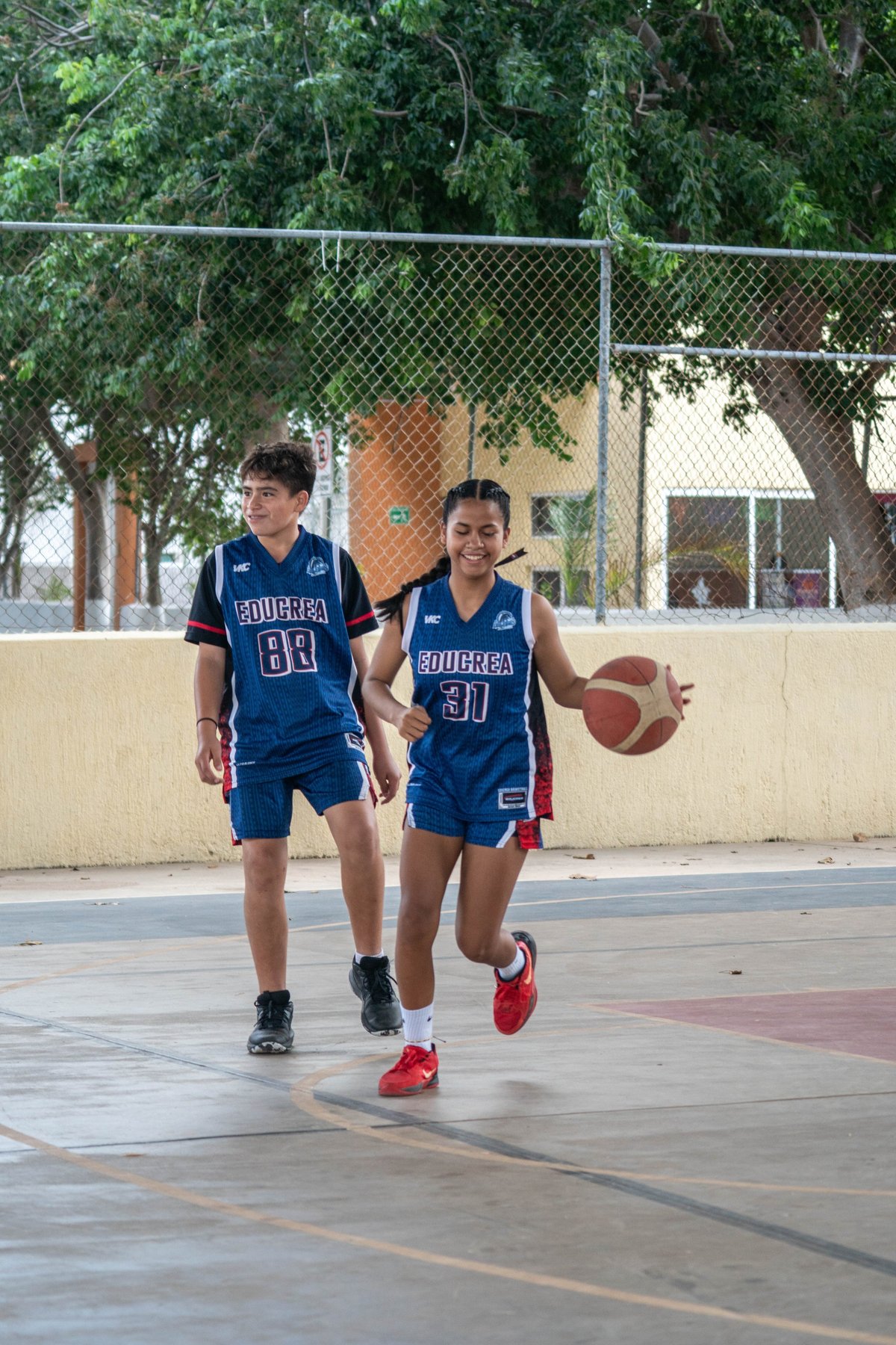 Dos jóvenes jugadores de básquetbol con uniformes azules Bourrea driblando en una cancha exterior con reja y árboles de fondo